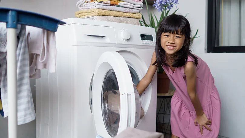 Imagen de una niña metiendo una manta en una lavadora.