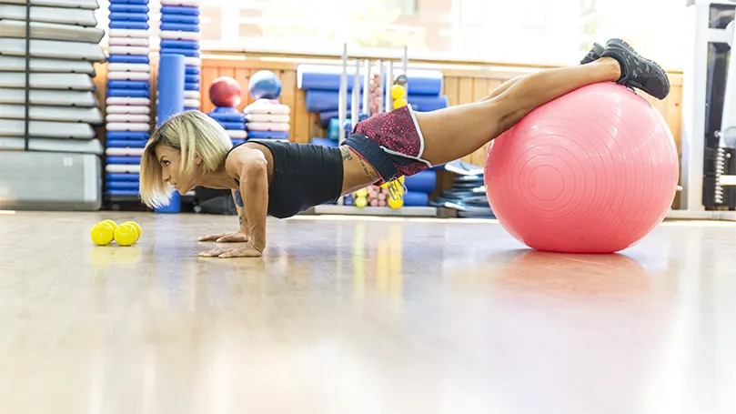 Una imagen de una mujer haciendo flexiones Decline sobre una pelota de yoga