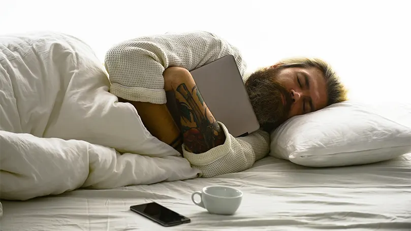 Imagen de un hombre con barba que duerme profundamente abrazado a una tableta.