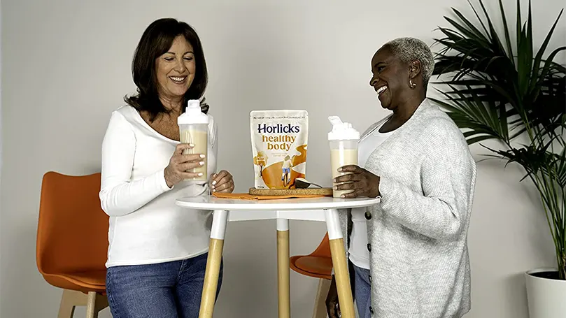 Dos mujeres sosteniendo vasos de horlicks y de pie junto a una mesa