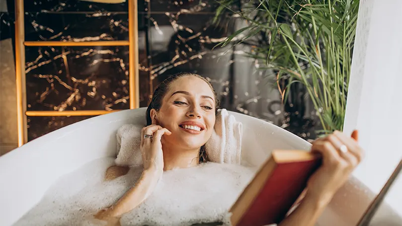 Una imagen de una mujer cómodamente tumbada en una bañera llena leyendo un libro y disfrutando.