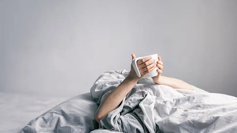 An image of a person covered with duvet holding a cup.