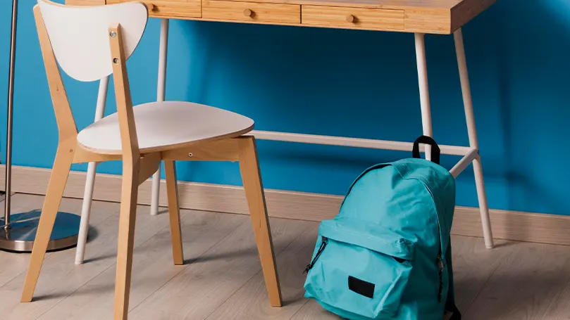 Pared azul, suelo de madera con un escritorio y una silla de madera. Una mochila azul colocada delante.